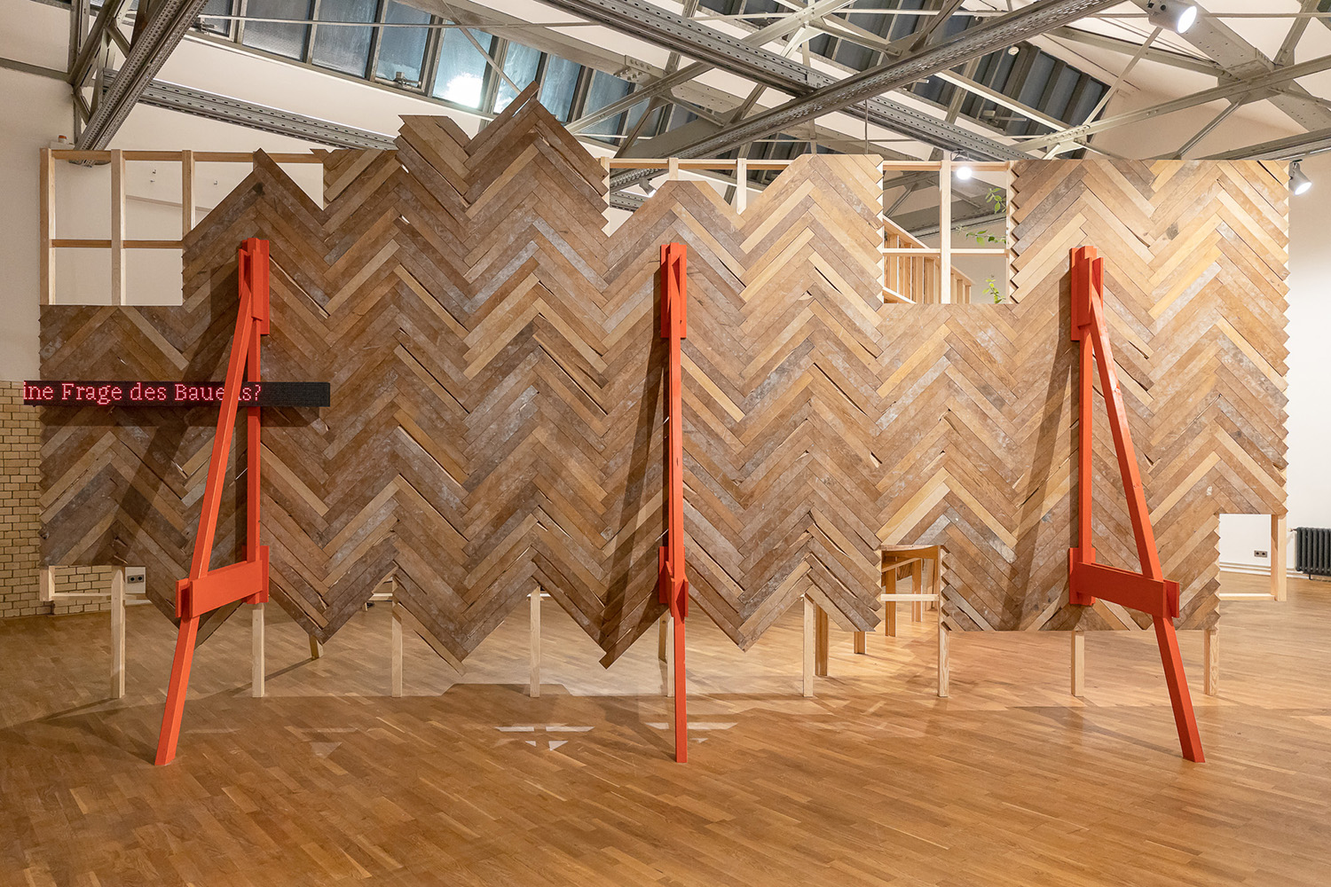 Foto einer ausgestellten Wand aus einem Holzgerüst, verkleidet mit Holzplatten im Fischgrätenmuster. Links an der Wand steht in digitaler Schrift " Eine Frage des Bauens?".